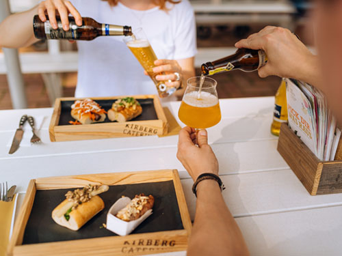 Zwei Personen gießen an einem Tisch im Freien Bier in Gläser, auf denen Essen auf Holztabletts steht.