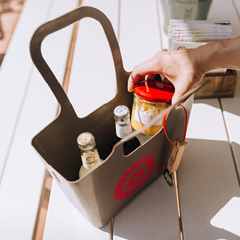 Eine Hand stellt ein Glas in eine graue Einkaufstasche mit Getränken auf einem weißen Tisch im Freien.