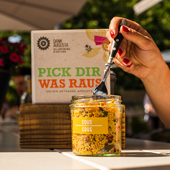 Eine Hand hält eine Gabel über ein Glas mit Couscous-Salat auf einem Tisch im Freien im Sonnenlicht.