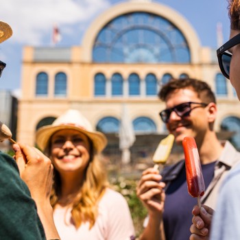Vier Freunde genießen an einem sonnigen Tag gemeinsam Eis am Stiel im Freien und lächeln vor einem großen, gewölbten Gebäude.