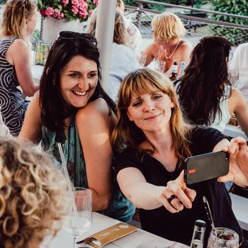 Zwei Frauen lächeln und posieren für ein Selfie in einem Straßenrestaurant mit anderen Menschen im Hintergrund.