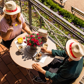 Zwei Personen mit Strohhüten genießen Getränke und Nachtisch an einem Cafétisch im Freien mit Blumenschmuck.