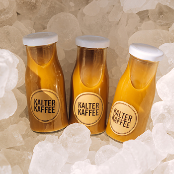 Drei Flaschen mit der Aufschrift „Kalter Kaffee“ stehen auf einem Bett aus Eiswürfeln.