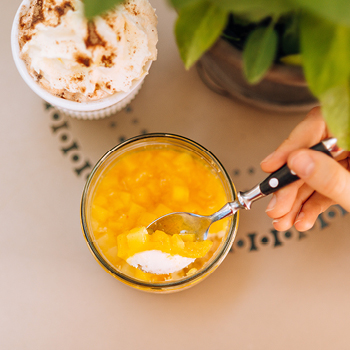Eine Hand hält einen Löffel über eine Glasschale mit Obst und Sahne; ein weiteres Dessert steht in der Nähe auf dem Tisch.