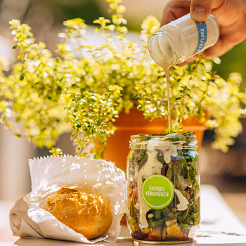 Eine Hand gießt Dressing in ein Glas Salat, daneben steht ein Brötchen, im Hintergrund eine Topfpflanze.