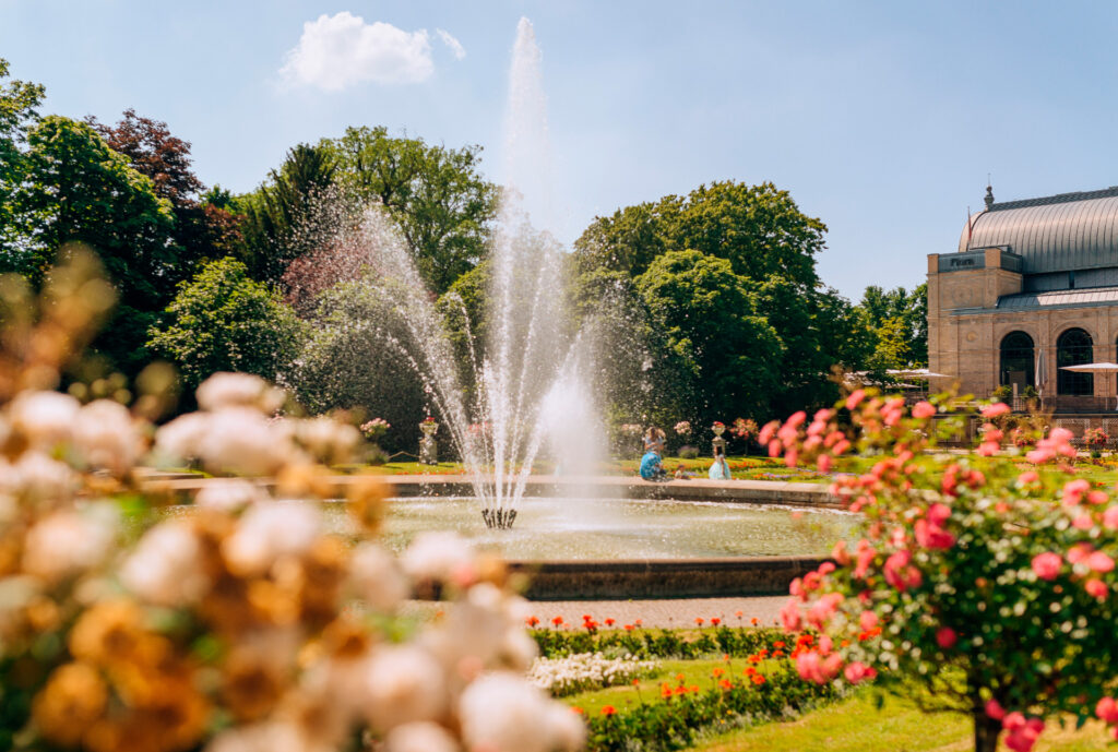 Ein großer Brunnen, umgeben von bunten Blumen und Grün in einem sonnigen Park mit einem historischen Gebäude in der Nähe.
